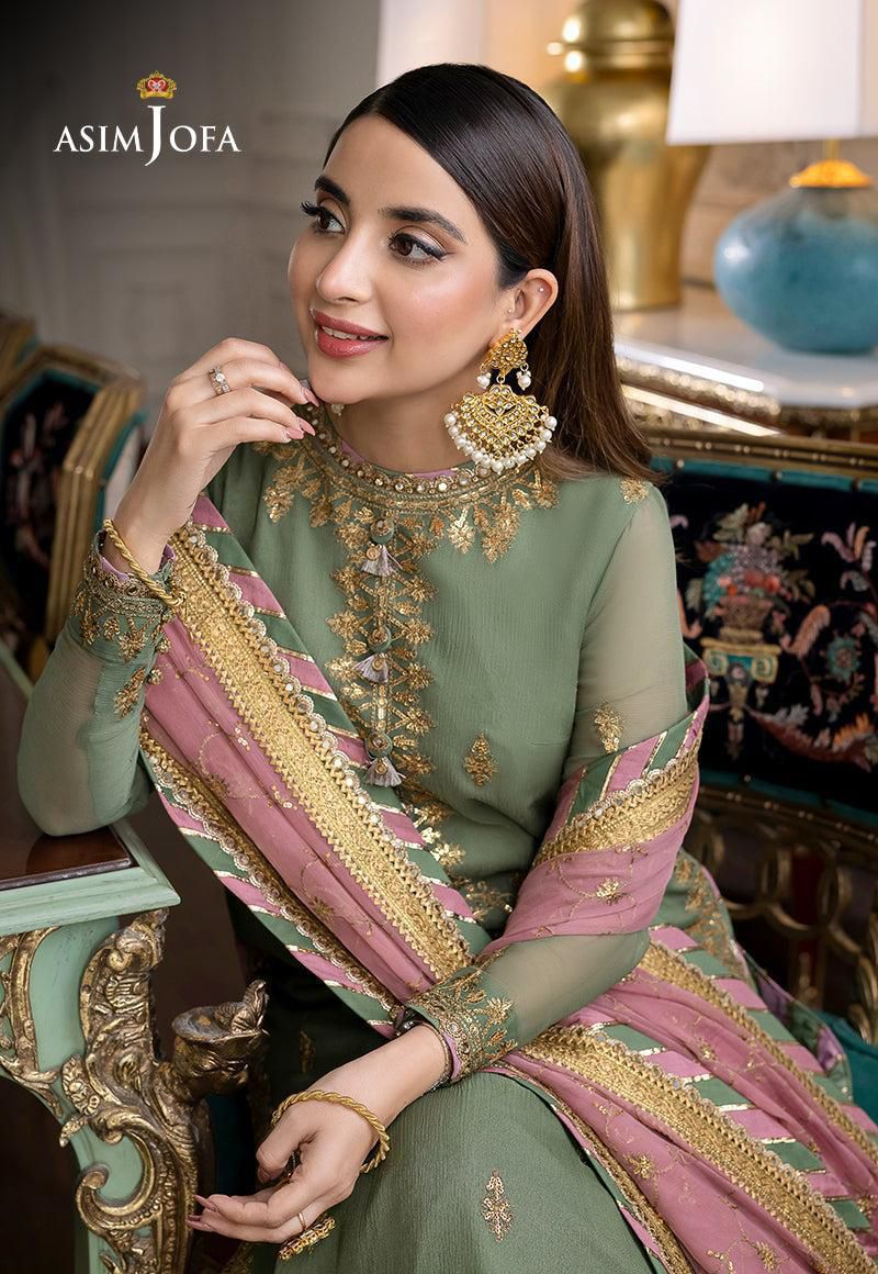 Woman wearing a traditional green and pink embroidered outfit with gold details, sitting in an elegant room.