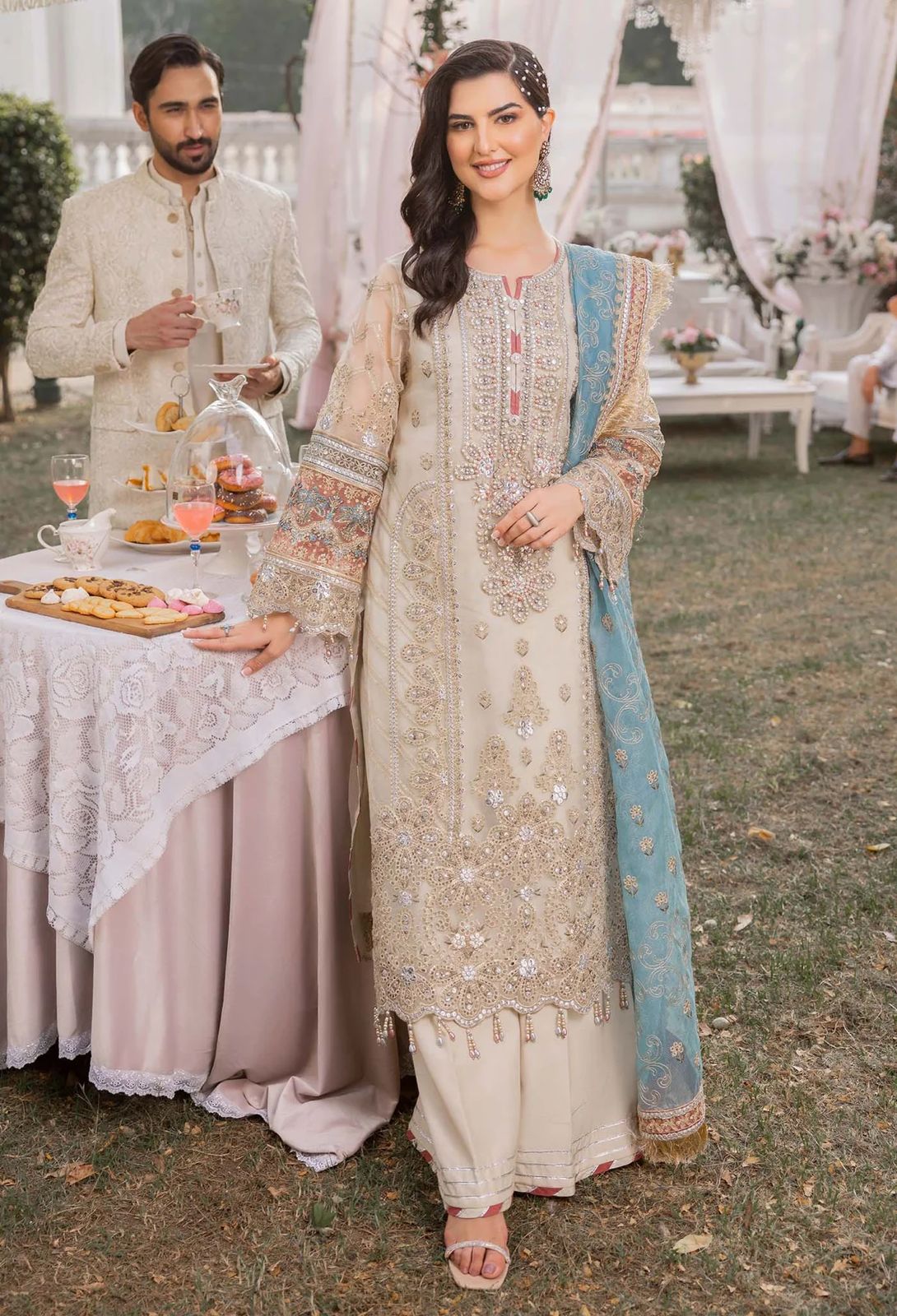 Woman in traditional embroidered outfit standing outdoors with a man and table in the background