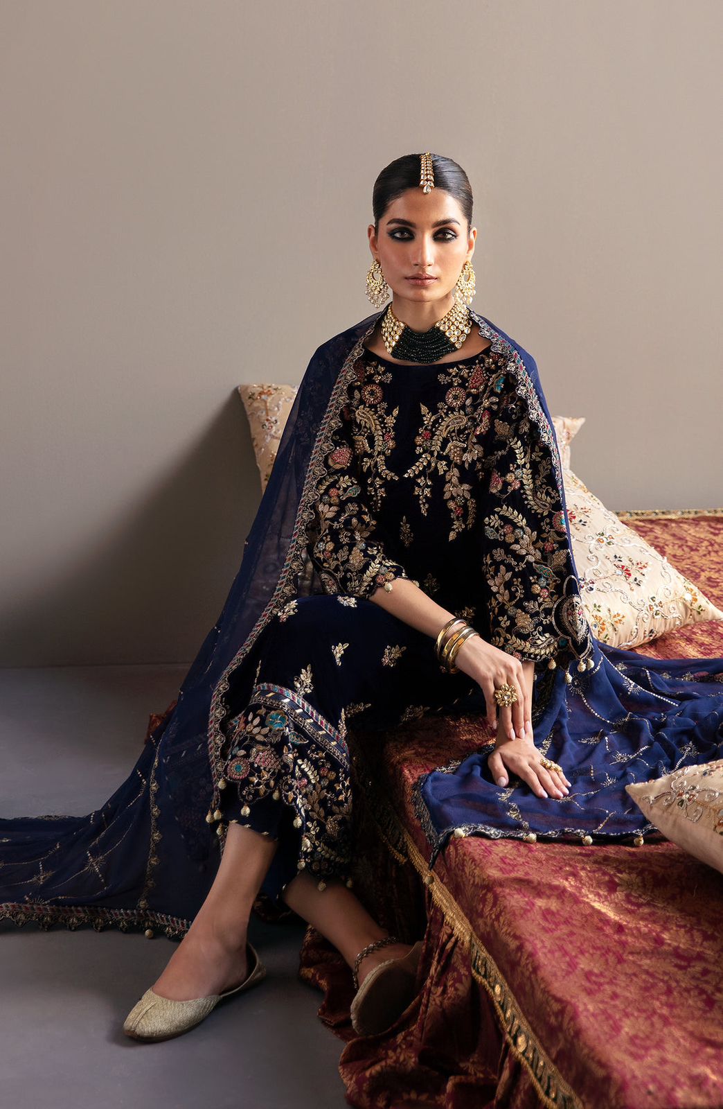 Emaan Adeel Velvet Woman in traditional attire sitting on a decorative couch against a plain background