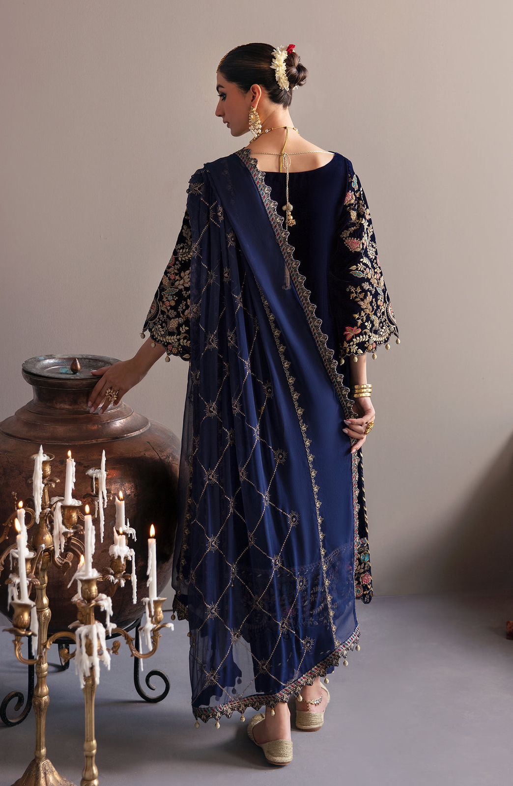Emaan Adeel Velvet Woman in a blue embroidered outfit standing next to a large vase and candelabra.