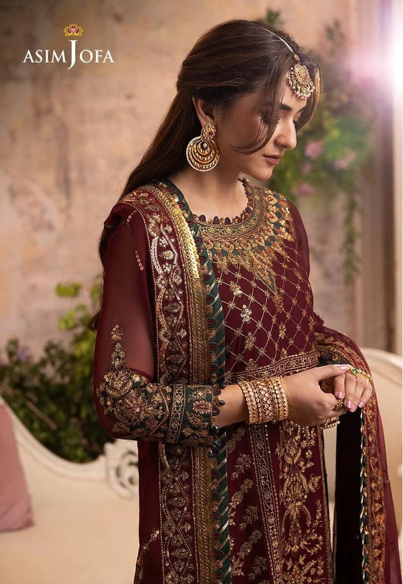Woman wearing an embroidered maroon traditional outfit with gold details, standing indoors.