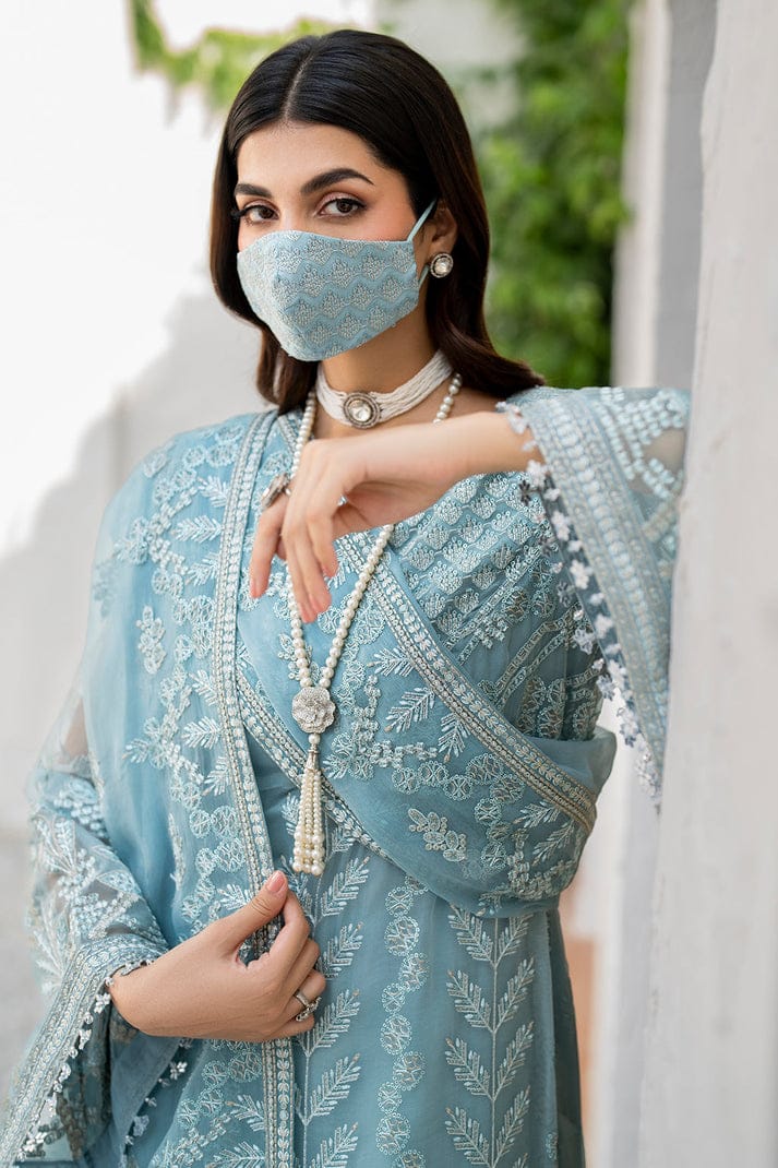 Flossie Blossom Woman wearing a light blue embroidered outfit with a matching face mask, standing against a white wall with greenery.