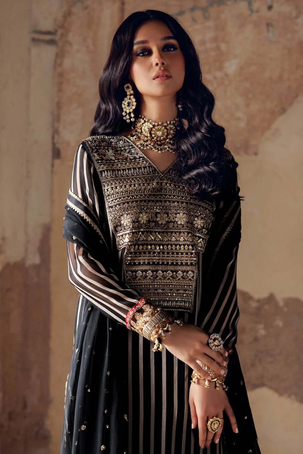 Charizma in traditional attire with gold jewelry against a textured wall.