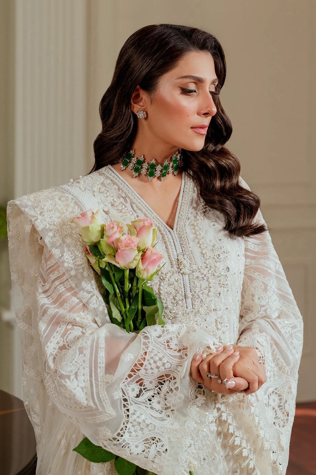 Woman in a white lace outfit holding pink roses indoors.