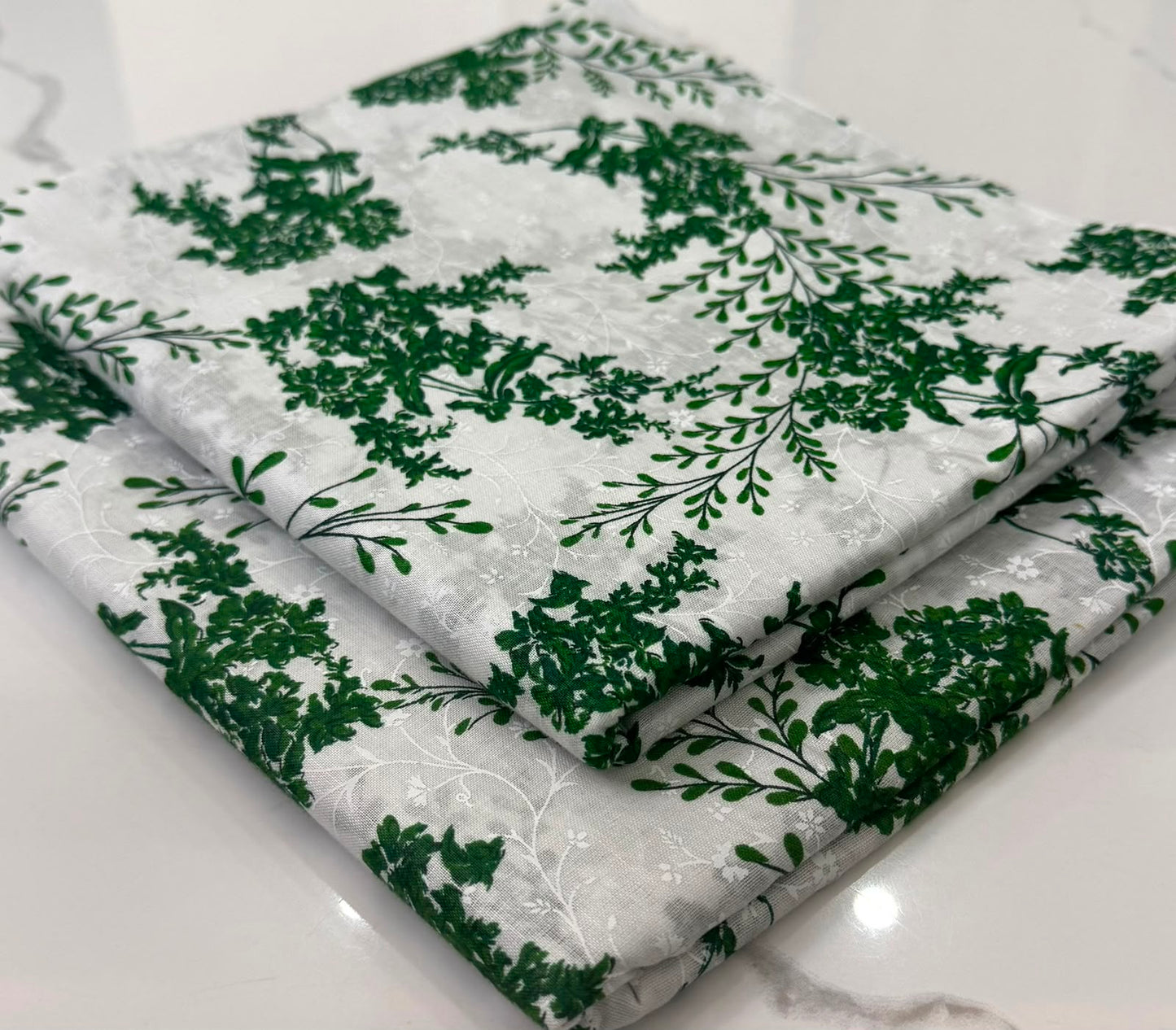 A folded piece of fabric with a green botanical leaf pattern on a white background, displayed on a light marble surface.