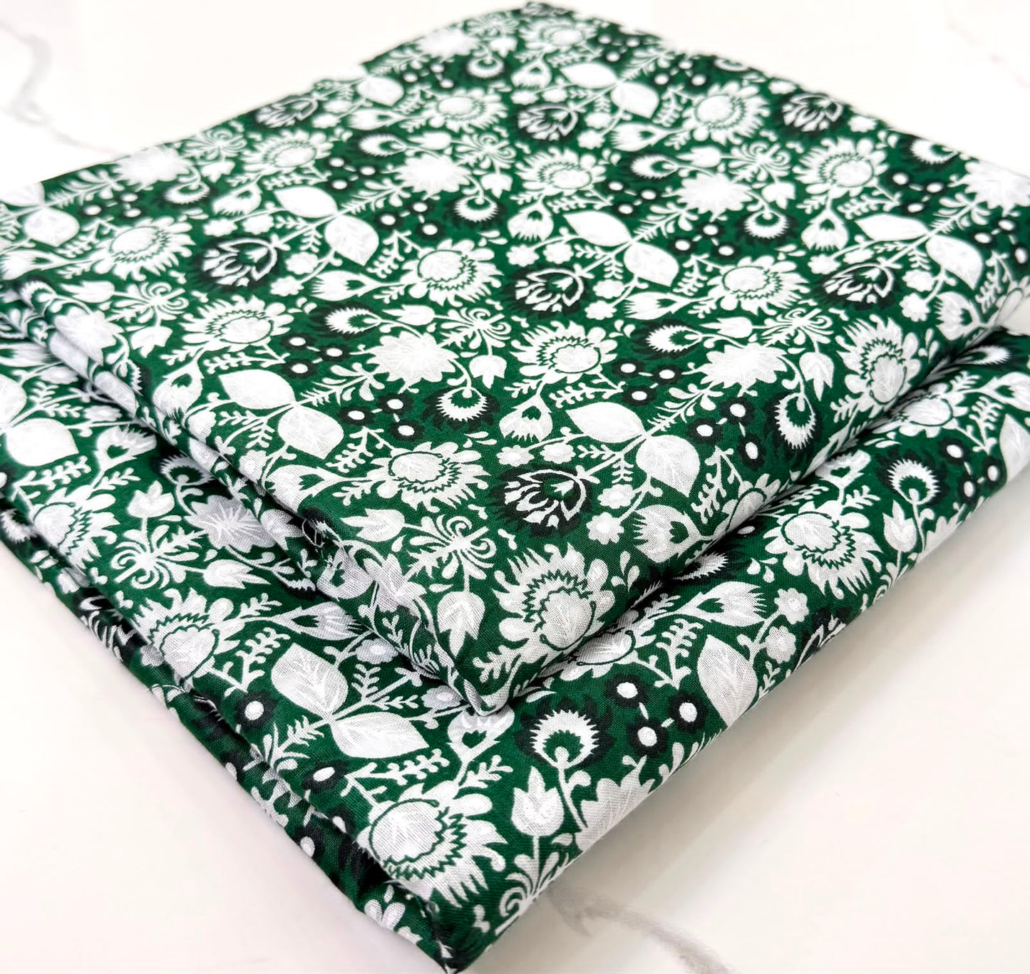 A folded piece of dark green fabric with a white floral and botanical pattern, displayed on a light marble surface.