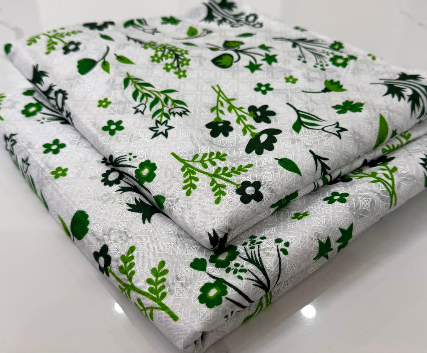 A folded white fabric item, likely a quilt or comforter, featuring a repeating pattern of green and black botanical leaves and flowers.