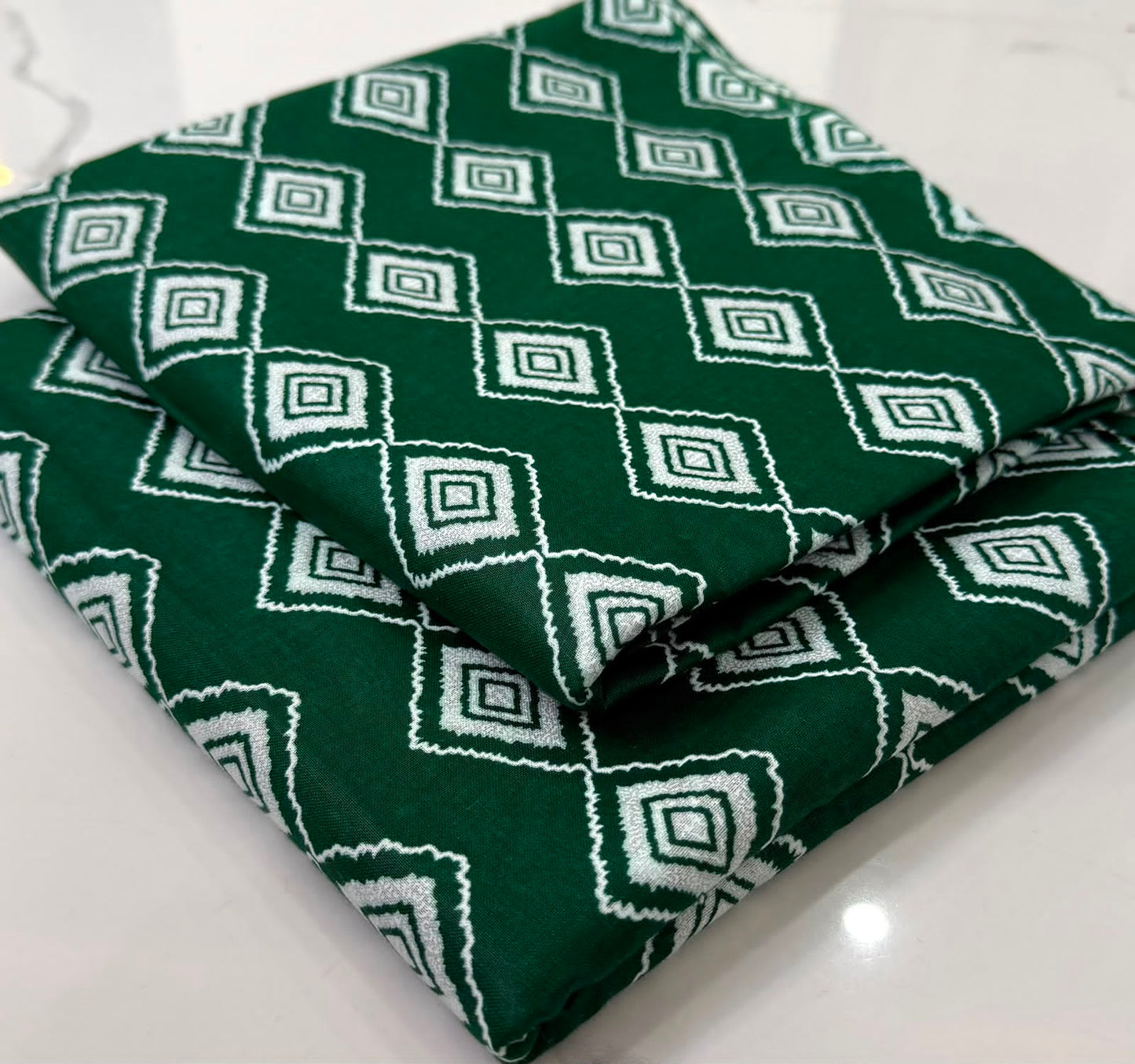 A folded dark green fabric with a white geometric diamond pattern, displayed on a light marble surface.