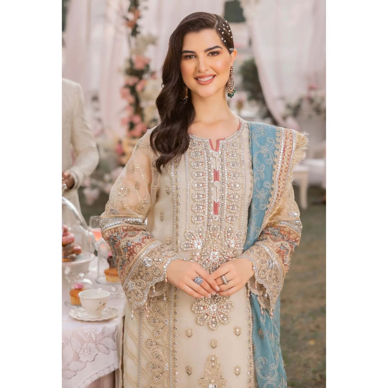 A woman wearing an ornate beige and light blue traditional embroidered outfit with intricate metallic detailing, photographed in an outdoor event setting with tables and draped fabric in the background.