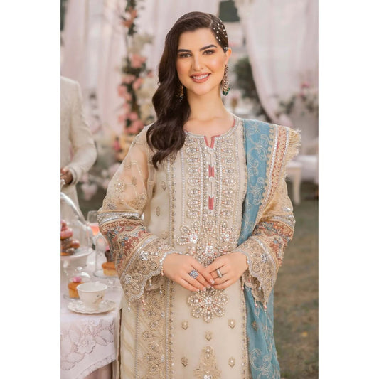 A woman wearing an ornate beige and light blue traditional embroidered outfit with intricate metallic detailing, photographed in an outdoor event setting with tables and draped fabric in the background.