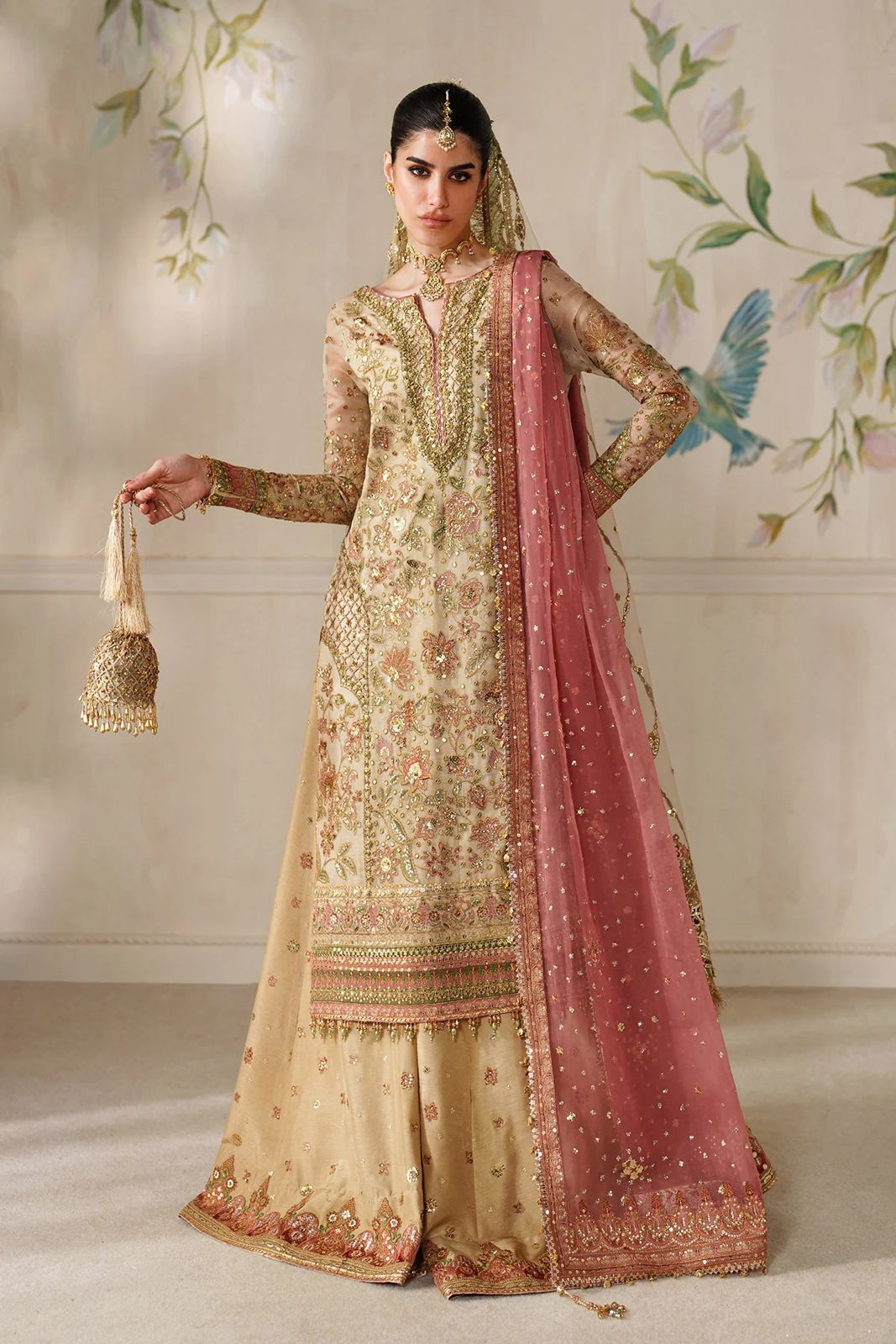Woman in traditional embroidered outfit with pink dupatta against a decorative wall.