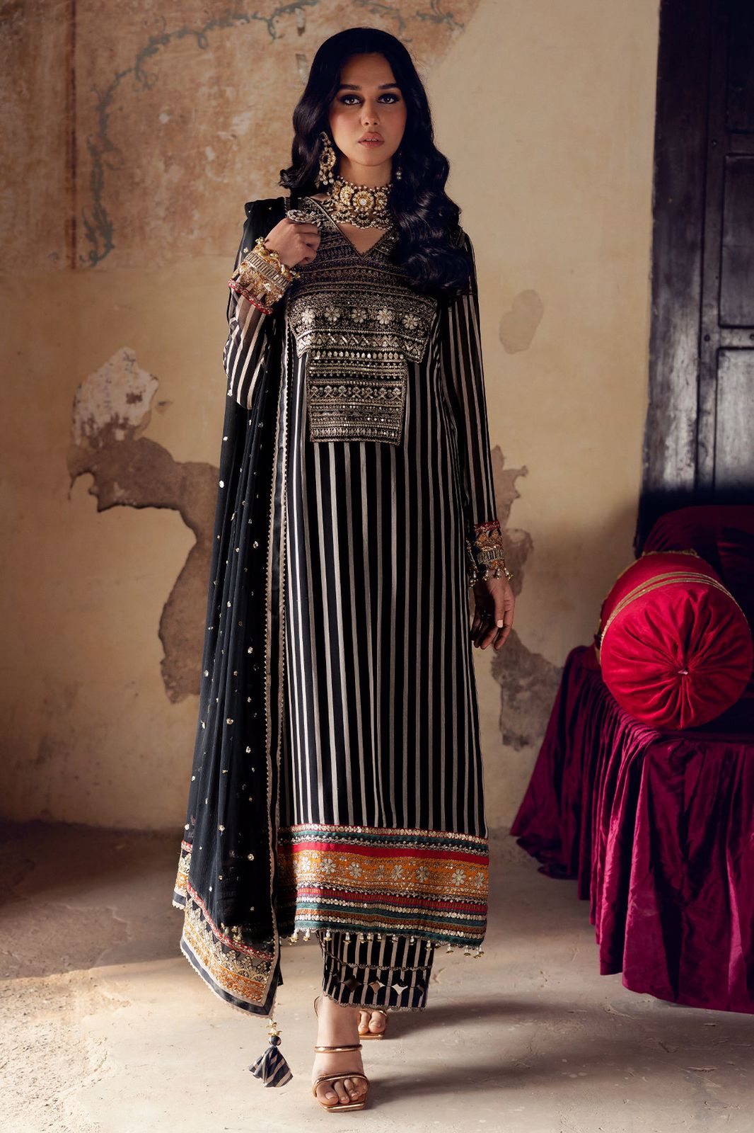 Woman in traditional black and gold outfit with intricate patterns in a room with a textured wall.