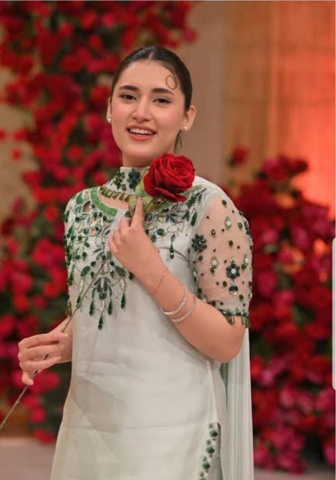A woman wearing a light green embroidered traditional outfit with sheer sleeves and holding a red rose, posed in front of a background of red flowers.