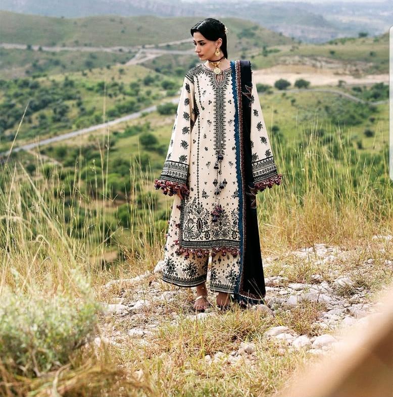 A woman stands outdoors in a grassy, hilly landscape wearing a traditional embroidered outfit with a cream base, black and navy border detailing, and multicolor floral motifs. The background shows rolling green hills and dry grass.
