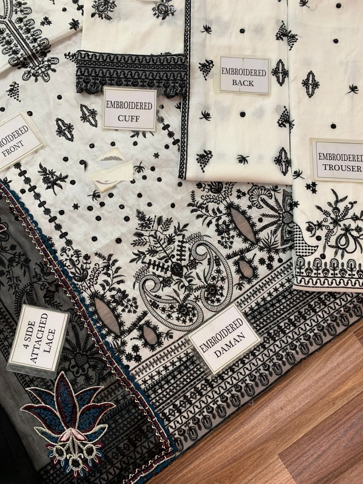 A folded embroidered fabric set laid out on a wooden floor, showing multiple pieces labeled with names such as embroidered cuff, embroidered back, embroidered trouser, embroidered front, and embroidered damask. The fabric is predominantly off-white with black floral and paisley embroidery, plus small blue and brown embroidered accents.