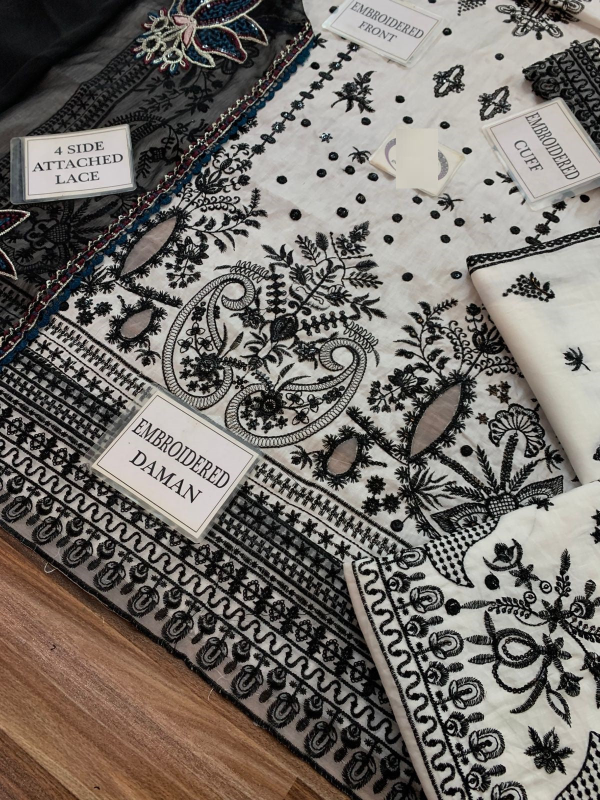 A folded embroidered fabric set with a white base and black paisley, floral, and geometric patterns, shown with labels indicating embroidery details like attached lace and daman.