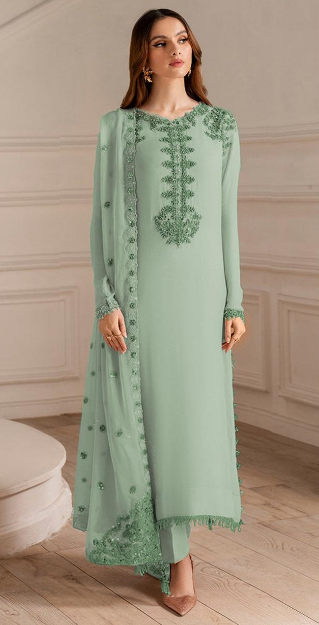 A woman models a mint green traditional Indian salwar kameez suit with intricate green floral embroidery on the chest and sleeves, paired with matching embroidered dupatta and trousers, set against a classic cream-colored paneled wall and wooden floor.