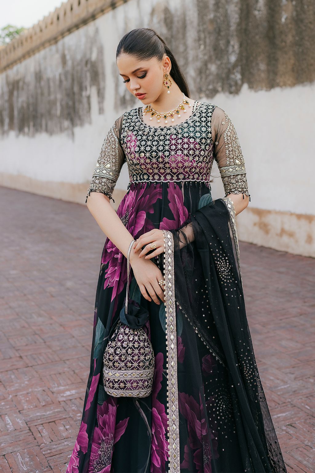 Woman in traditional outfit with floral patterns and jewelry, holding a matching handbag.