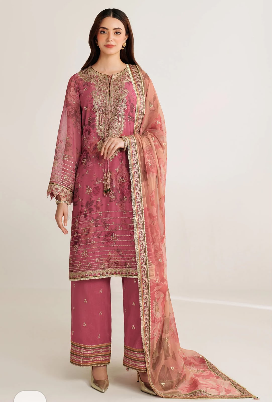 A woman modeling a traditional three-piece salwar kameez suit in dusty pink with intricate gold embroidery, paired with matching trousers and a sheer pink dupatta with a gold border, set against a plain light gray studio background.