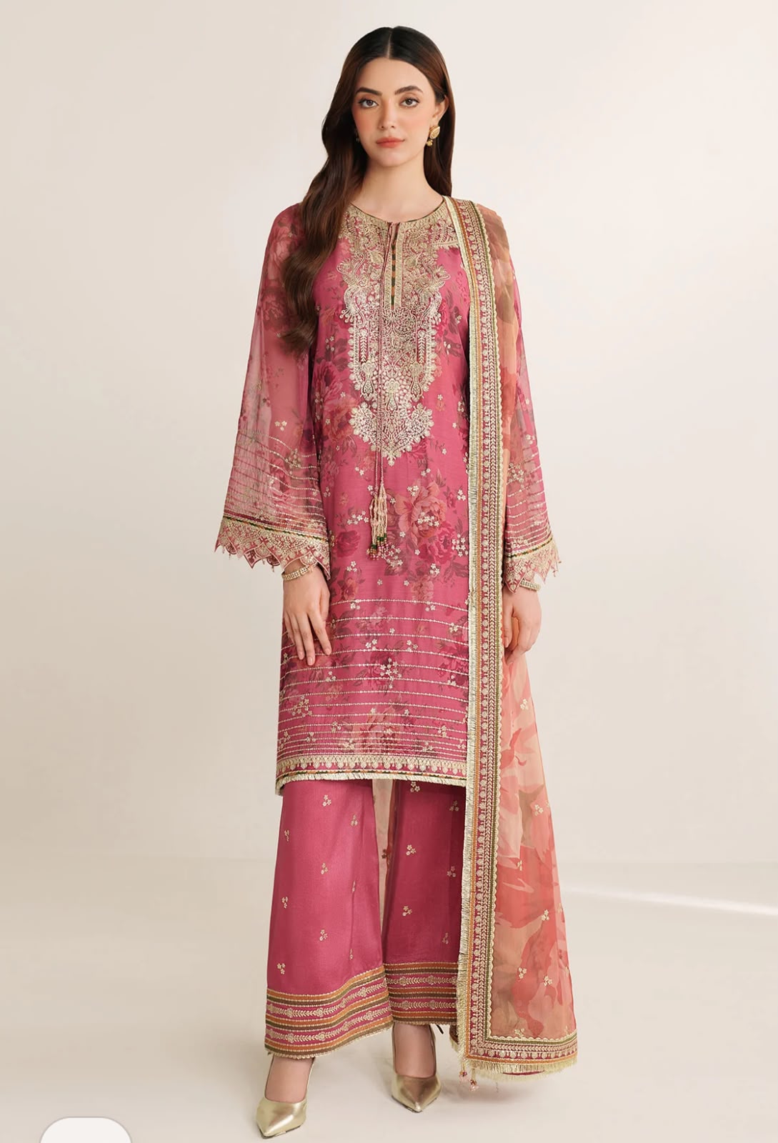 A woman modeling a pink traditional three-piece outfit with gold embroidery and a matching sheer dupatta, photographed against a plain light beige studio background.
