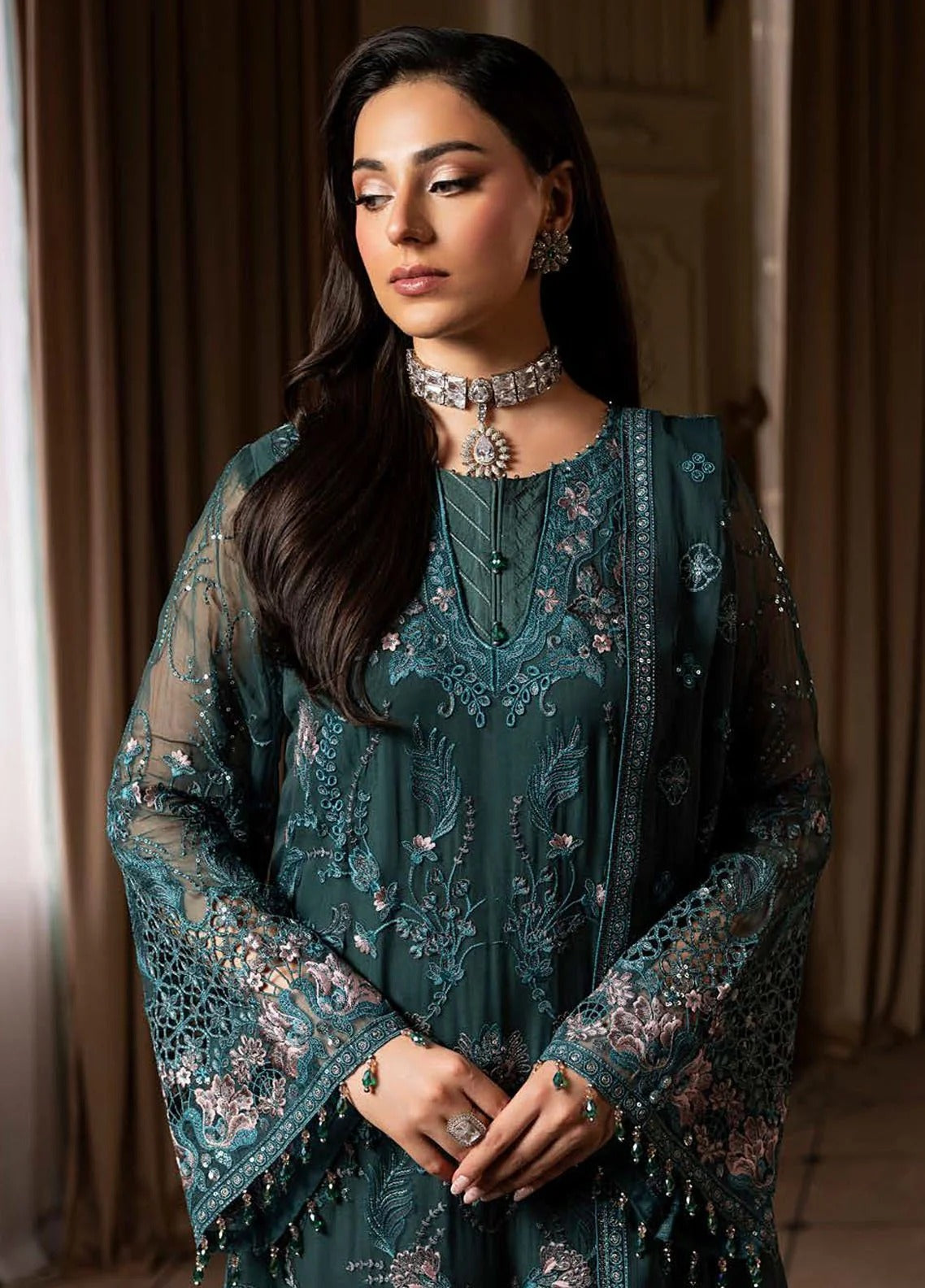 Woman wearing a teal embroidered outfit with jewelry indoors.