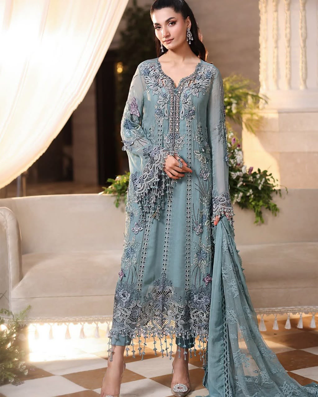 A woman models a traditional long teal blue ethnic suit with intricate silver and gold embroidery, a matching dupatta, and silver heels in an elegant indoor setting.