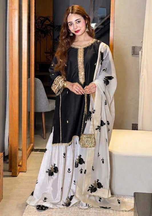 A woman models a traditional black and white embroidered kurta set with a matching dupatta, featuring gold embroidery and black floral prints, in an indoor setting.