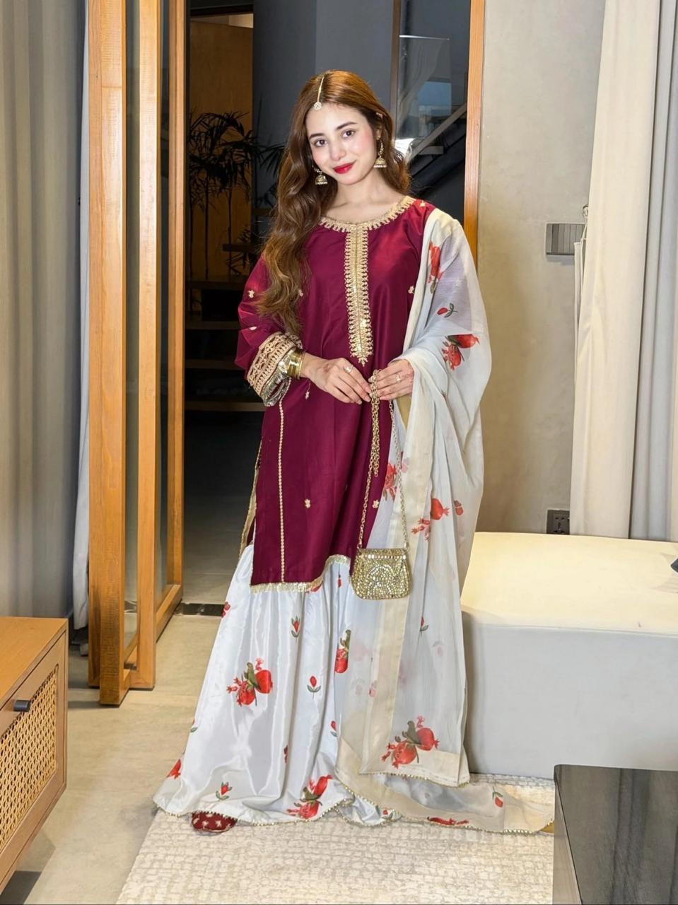 A woman models a traditional South Asian outfit consisting of a deep maroon kurta with gold embroidery, matching flared pants, and a sheer white dupatta with red floral prints, standing in an indoor setting.