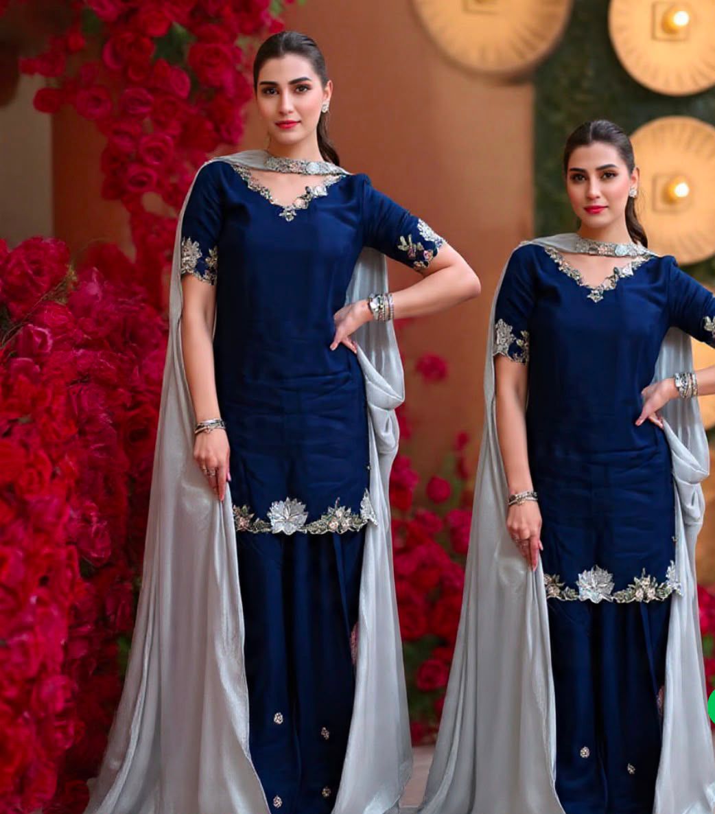 Two women modeling matching navy blue traditional outfits with silver floral embroidery and light gray dupattas, posed in front of a warm-toned backdrop with red floral arrangements.