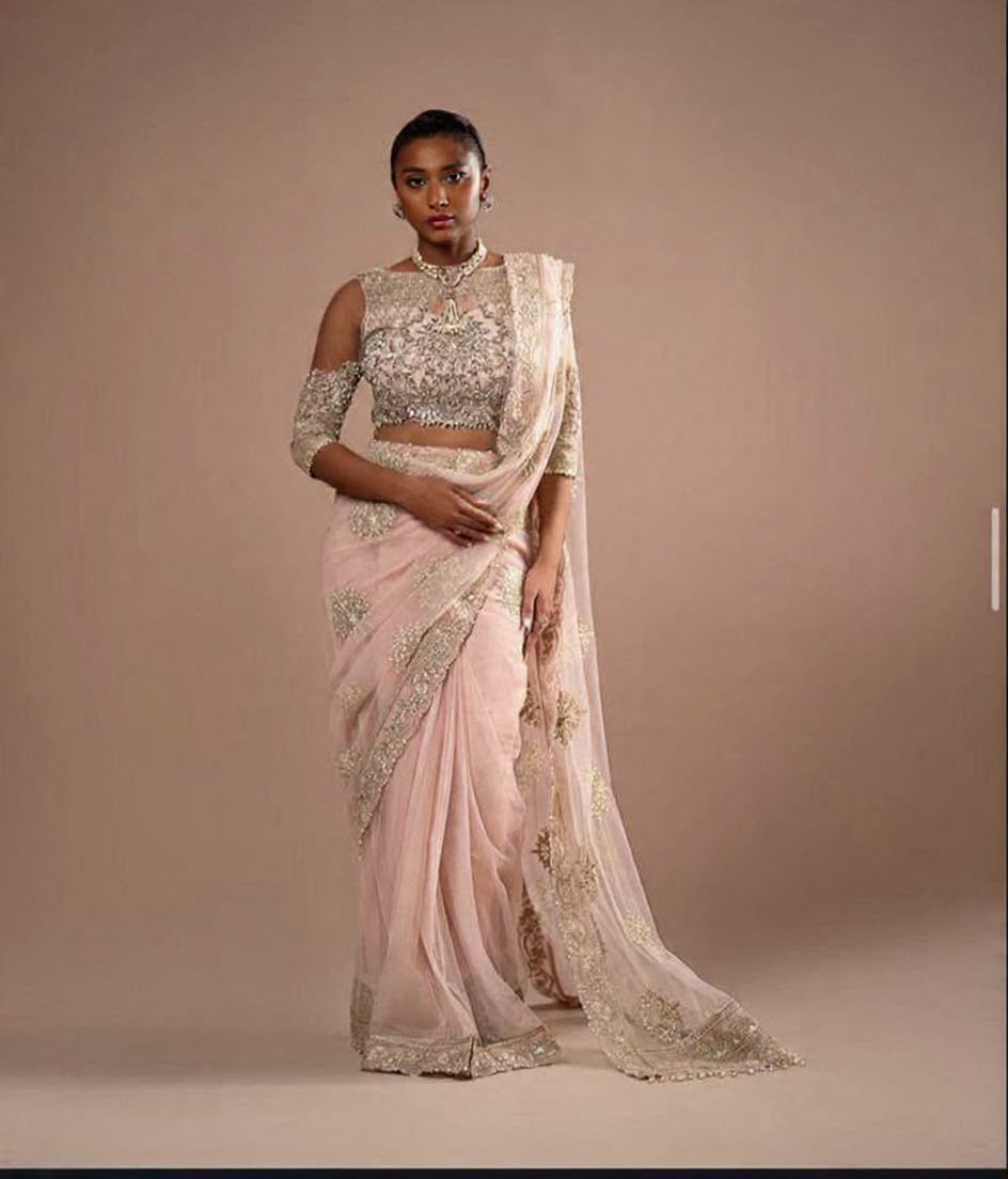 A woman modeling a light pink saree with gold embroidery and a matching embellished blouse against a plain beige studio background.
