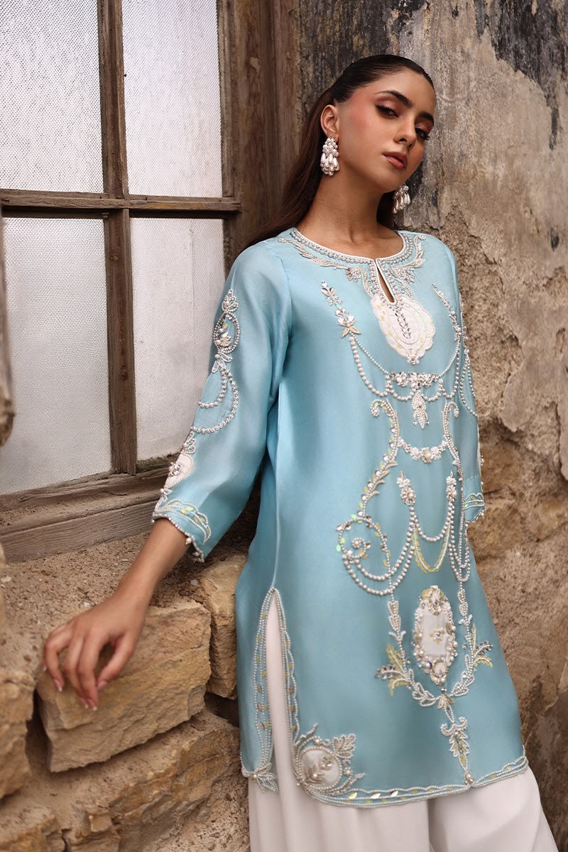 Woman wearing a light blue embroidered top with white pants against a rustic stone wall.
