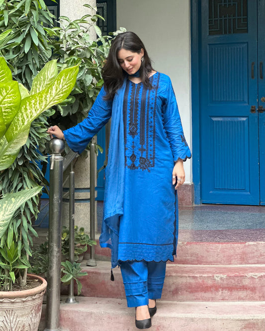 Woman in a blue traditional outfit standing on a staircase with plants and a blue door in the background.