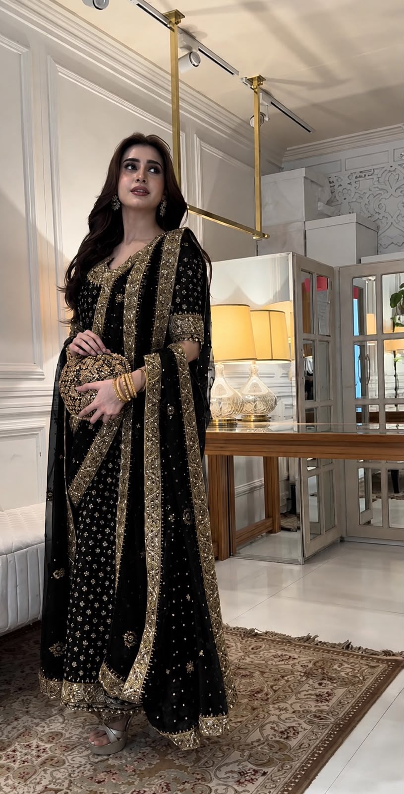 A woman models a black traditional outfit with extensive gold embroidery in an elegant indoor setting with a mirror, table, and warm lighting.